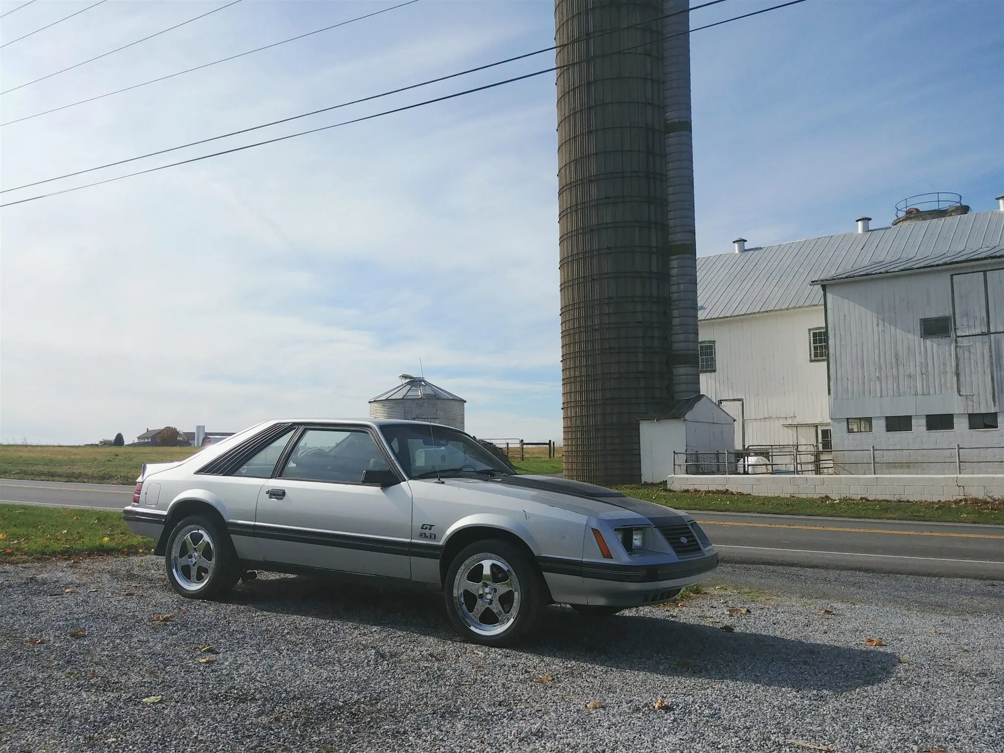 A silver 1986 Ford Mustang GT parked on a farm lane beside a silo